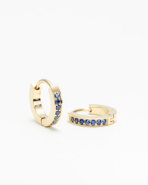 Pair of gold huggie hoop earrings set with a row of blue gemstones, laid on a white background.