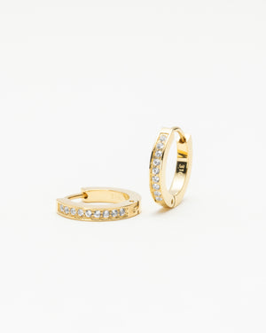 Pair of gold huggie hoop earrings set with a row of clear gemstones, laid on a white background.