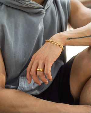 Man wearing gold chain-link bracelet with textured bar detail and gold band ring, seated with tattooed arm