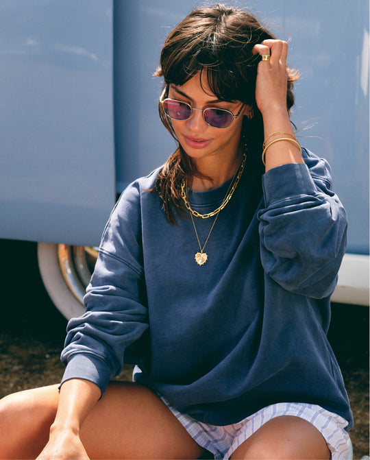 Woman in sunglasses and a blue sweatshirt wearing layered gold necklaces with a heart pendant.